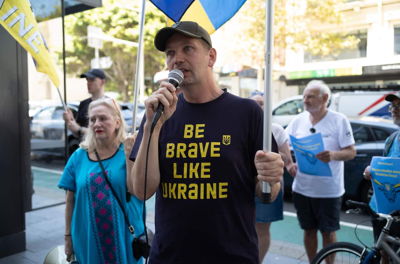 A man speaks at a rally into a microphone, his shirt reads 'Be brave like Ukraine'