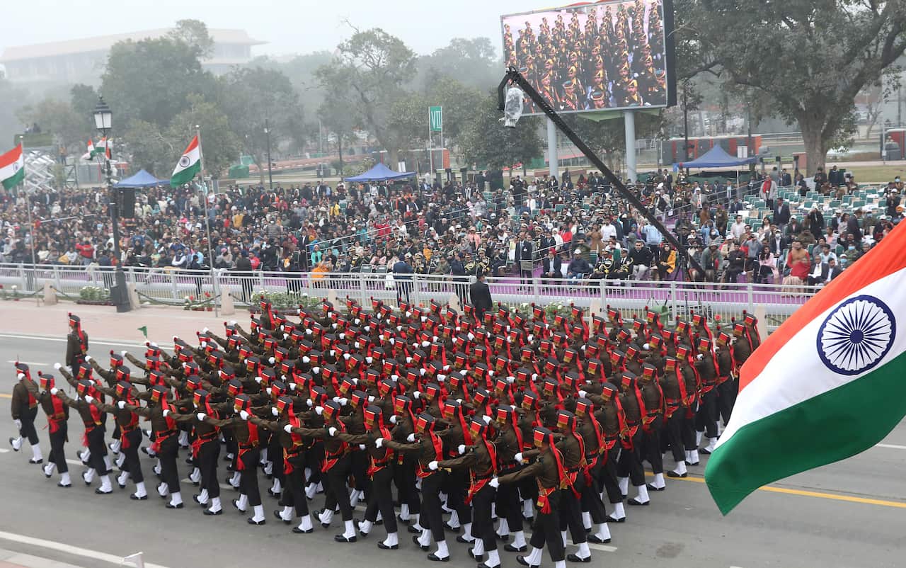 An army unit parades down a street with India's flag visible in the foreground.