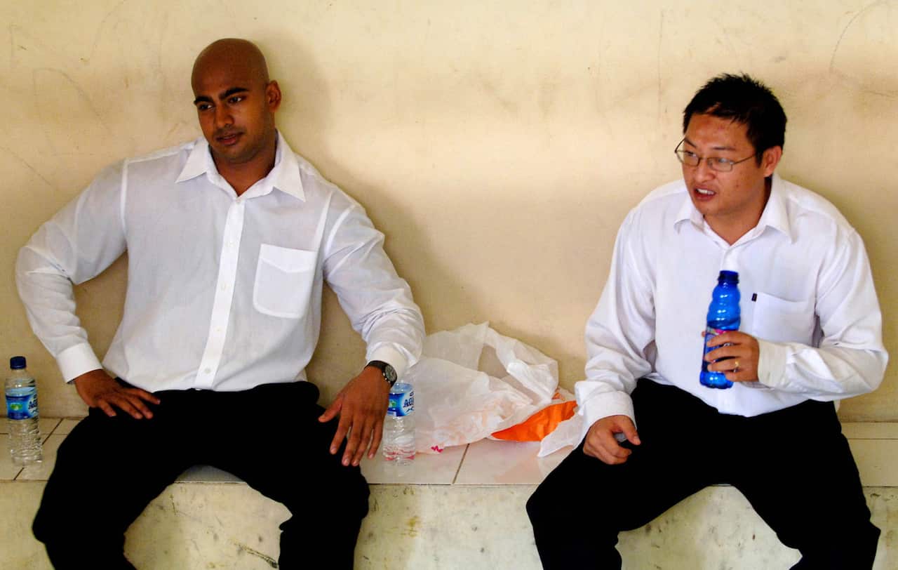 Two persons wearing white shirts and black pants sitting in a holding cell.