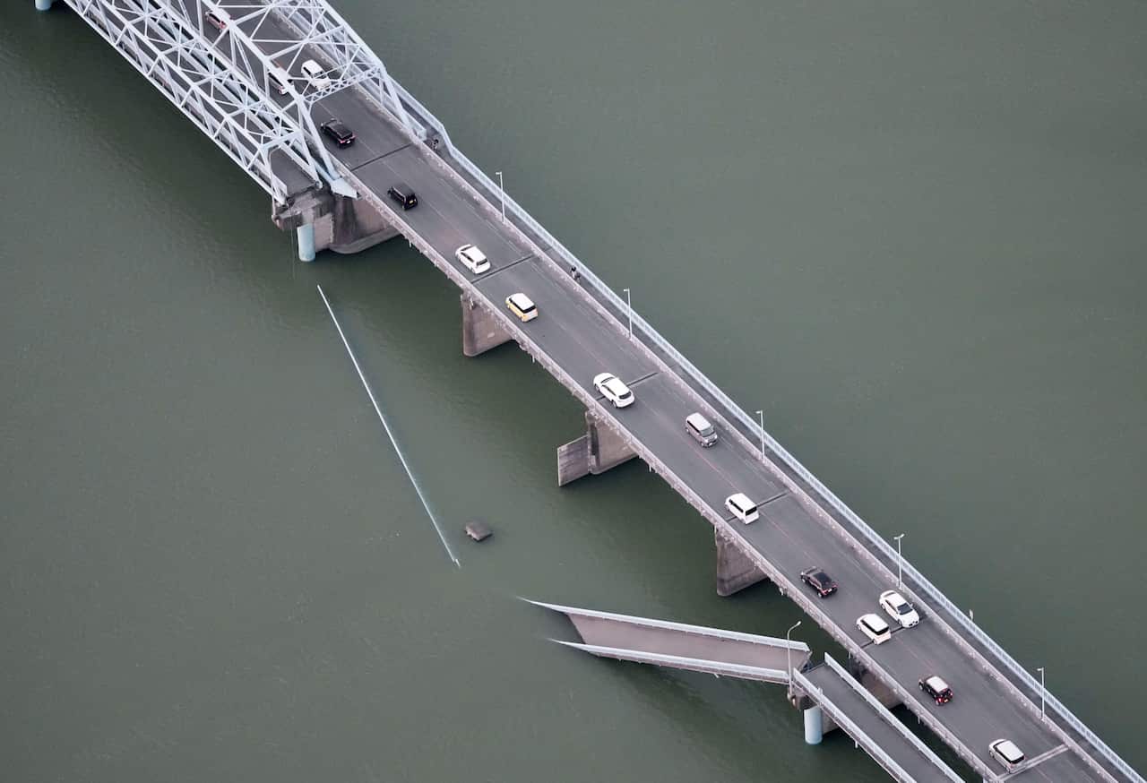 A partially collapsed footbridge sits next to a two-lane road bridge over a body of water.