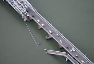 A partially collapsed footbridge sits next to a two-lane road bridge over a body of water.