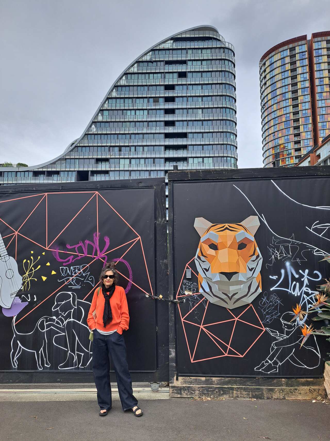 A woman wearing a bright red sweater stands in front of a large graffitied gate against a backdrop of high rise buildings. 
