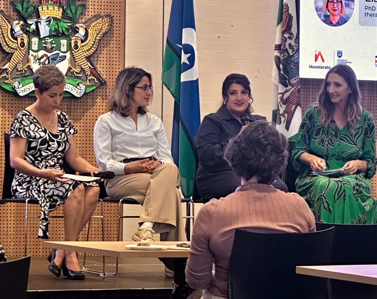 Four women sit on chairs in a half-circle arrangement on a raised platform while another woman with her back turned sits in front of them.
