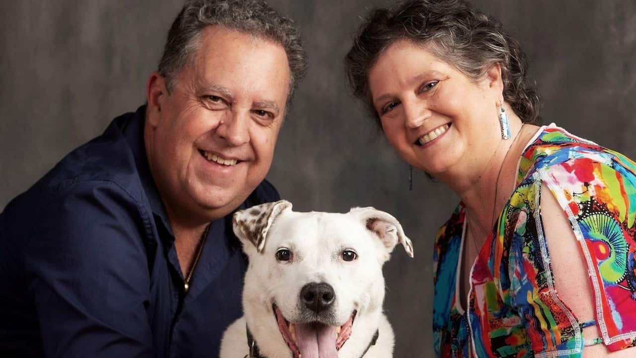 a man wearing a black shirt and a woman wearing a colourful shirt pose together for a photo with their white dog in the middle