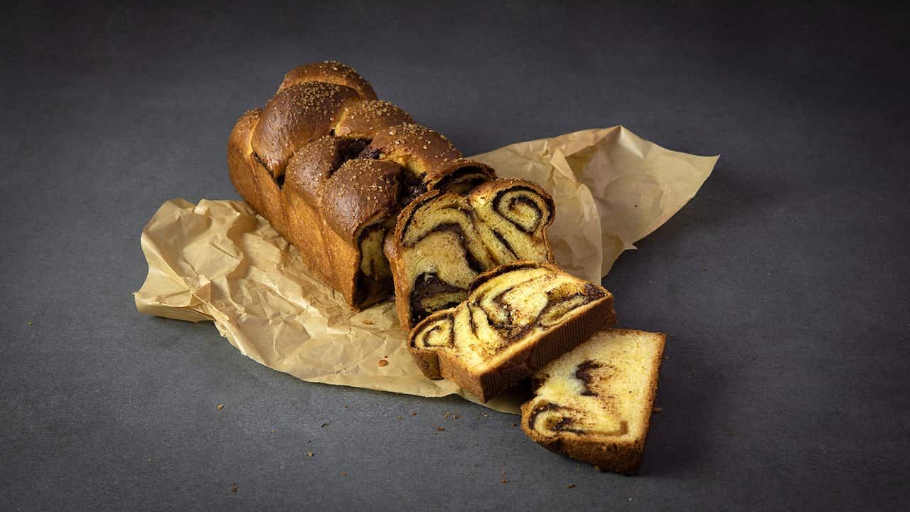 A loaf of golden plaited sweet bread sits on a piece of paper on a grey surface. The loaf has several slices cut from it, showing a swirl of chocolate running through the interior. 