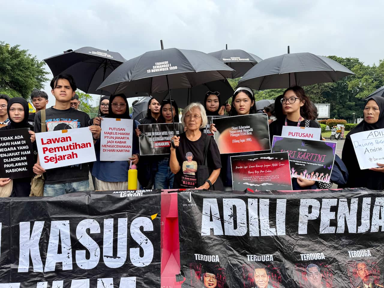 A group of people holding posters and a long banner are protesting. Some are holding black umbrellas.