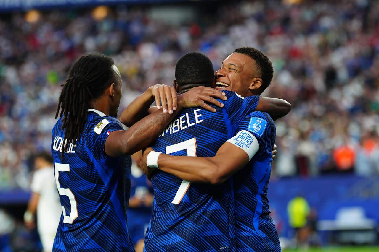 A footballer wearing a blue jersey smiles as he hugs his two teammates.