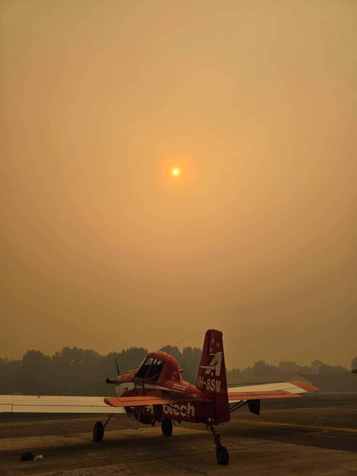 A red Fire Boss plane sits on the tarmac with the eerie orange haze of smoke covering the sky.