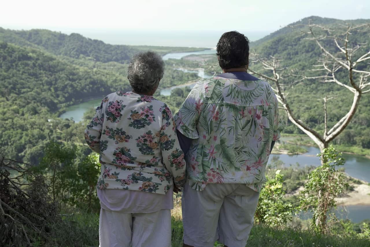 Two older women in print-pattern shirts stand side by side on a hilltop. Their backs are to the camera and they are overlooking a rainforest river running to the sea.