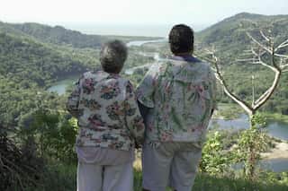 Two older women in print-pattern shirts stand side by side on a hilltop. Their backs are to the camera and they are overlooking a rainforest river running to the sea.