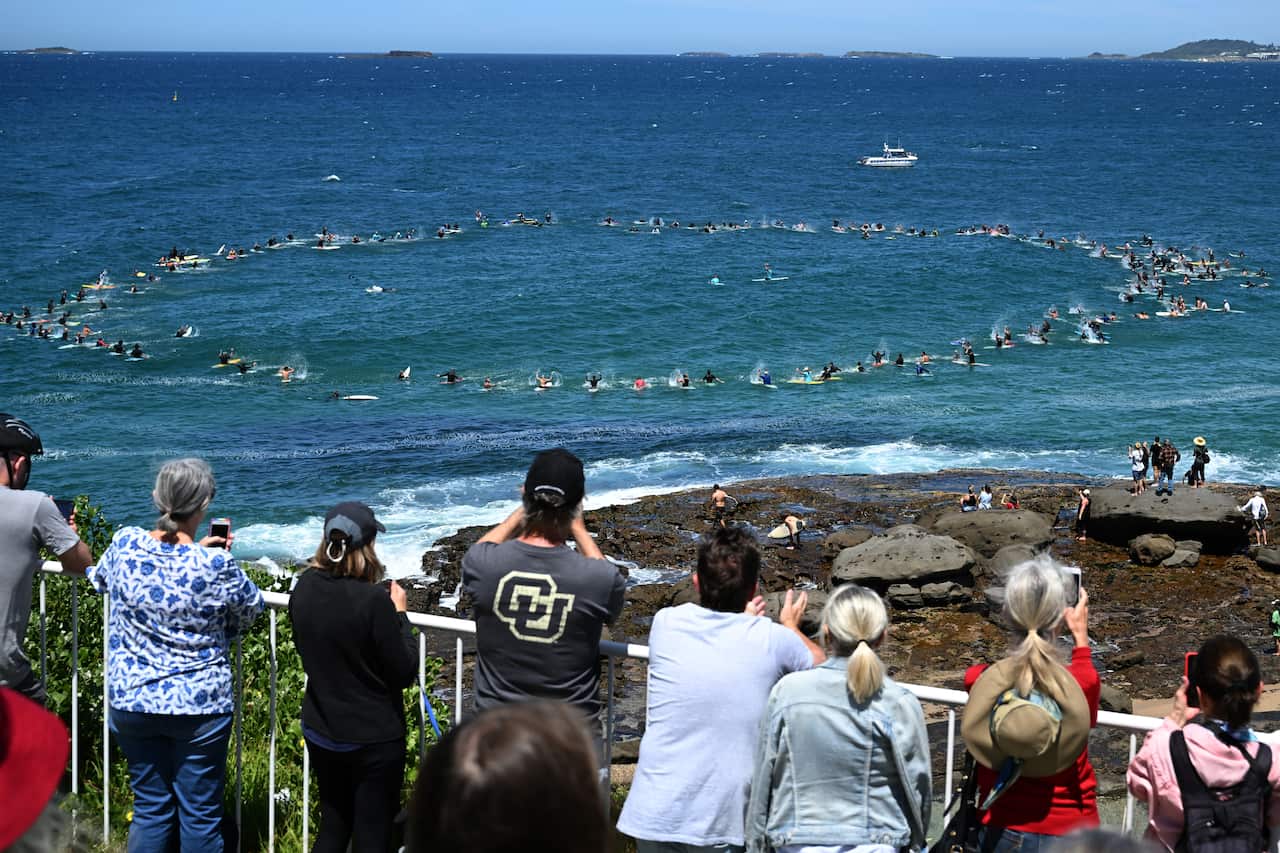 Surfers siting on boards in a circle in the ocean while a crowd on the shore watches