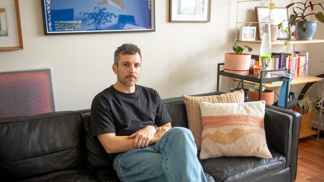 A man in a black t shirt and lightwash jeans sits on a black leather couch. He's looking at the camera with a neutral facial expression. There is art on the walls behind him, and a shelf with pictures and plants to his right.