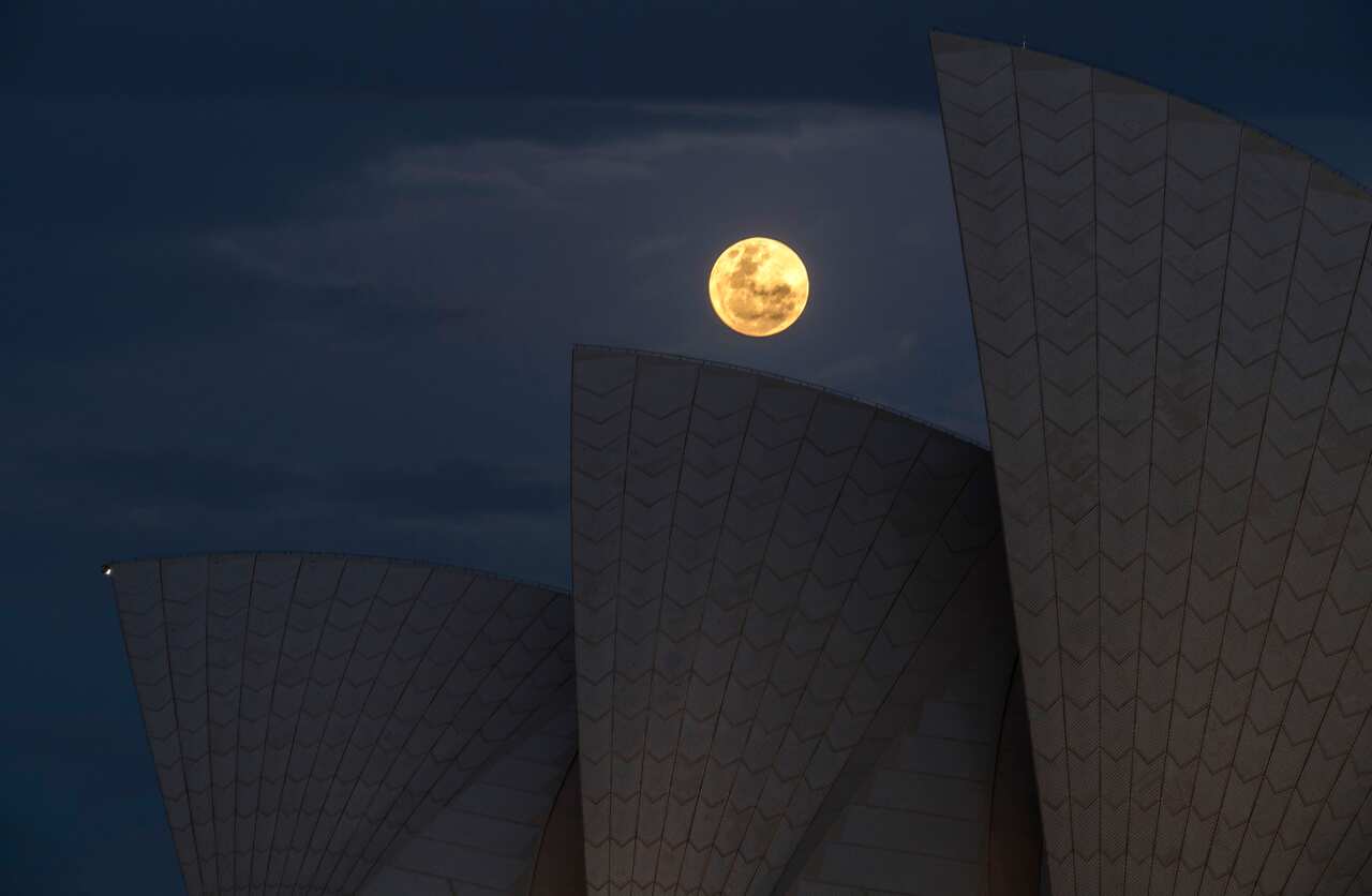 A bright, round supermoon illuminates the night sky above a sail-like structure.