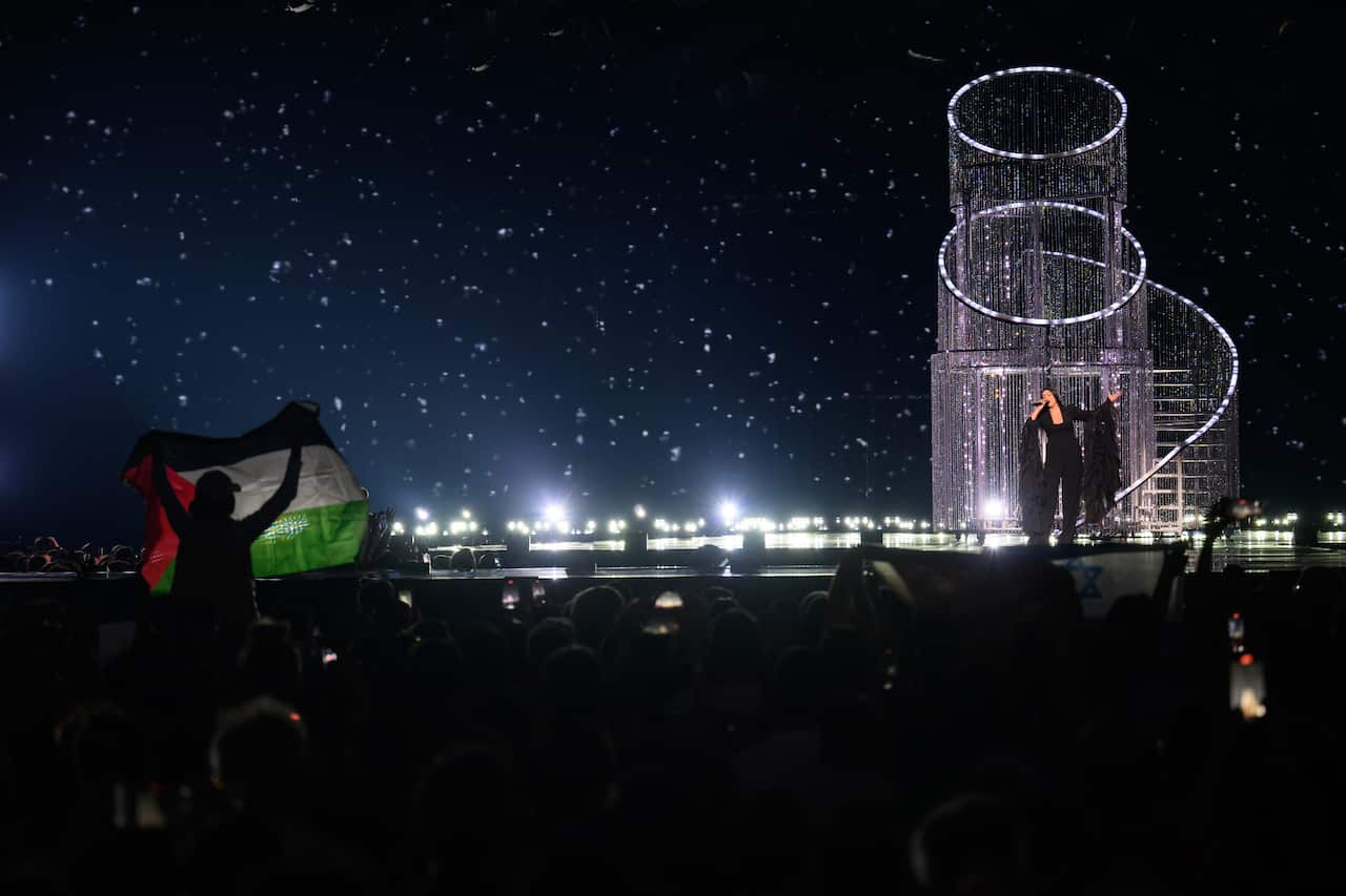 A singer performing on stage. A person is waving a Palestinian flag in the audience.