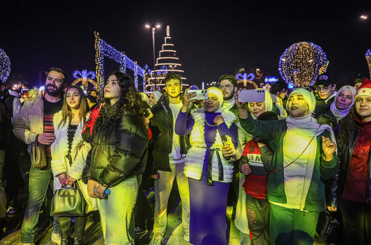 A group of people stand in front of the glow of artificial light. Behind them are several large objects decorated with Christmas lights. 