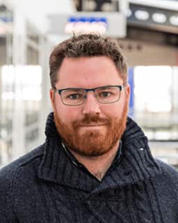 A man in glasses and a beard is wearing a woollen jumper as he poses.