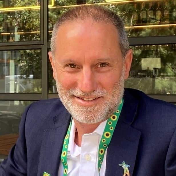 A man in a dark blue jacket and white shirt, with a yellow and green sunflower lanyard around his neck