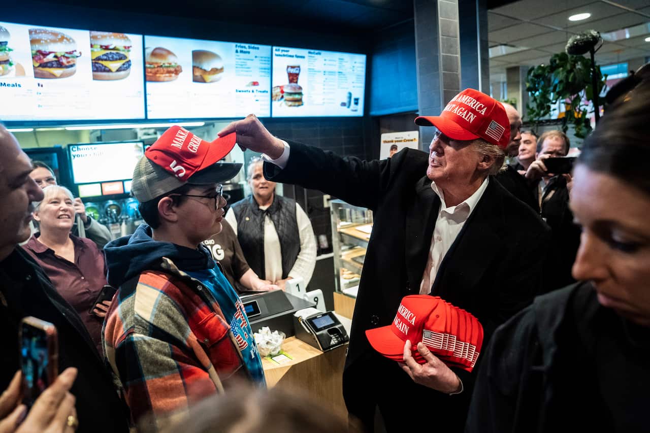 Donald Trump, wearing a red cap, is placing one on a young boy's head while holding several more as onlookers watch.