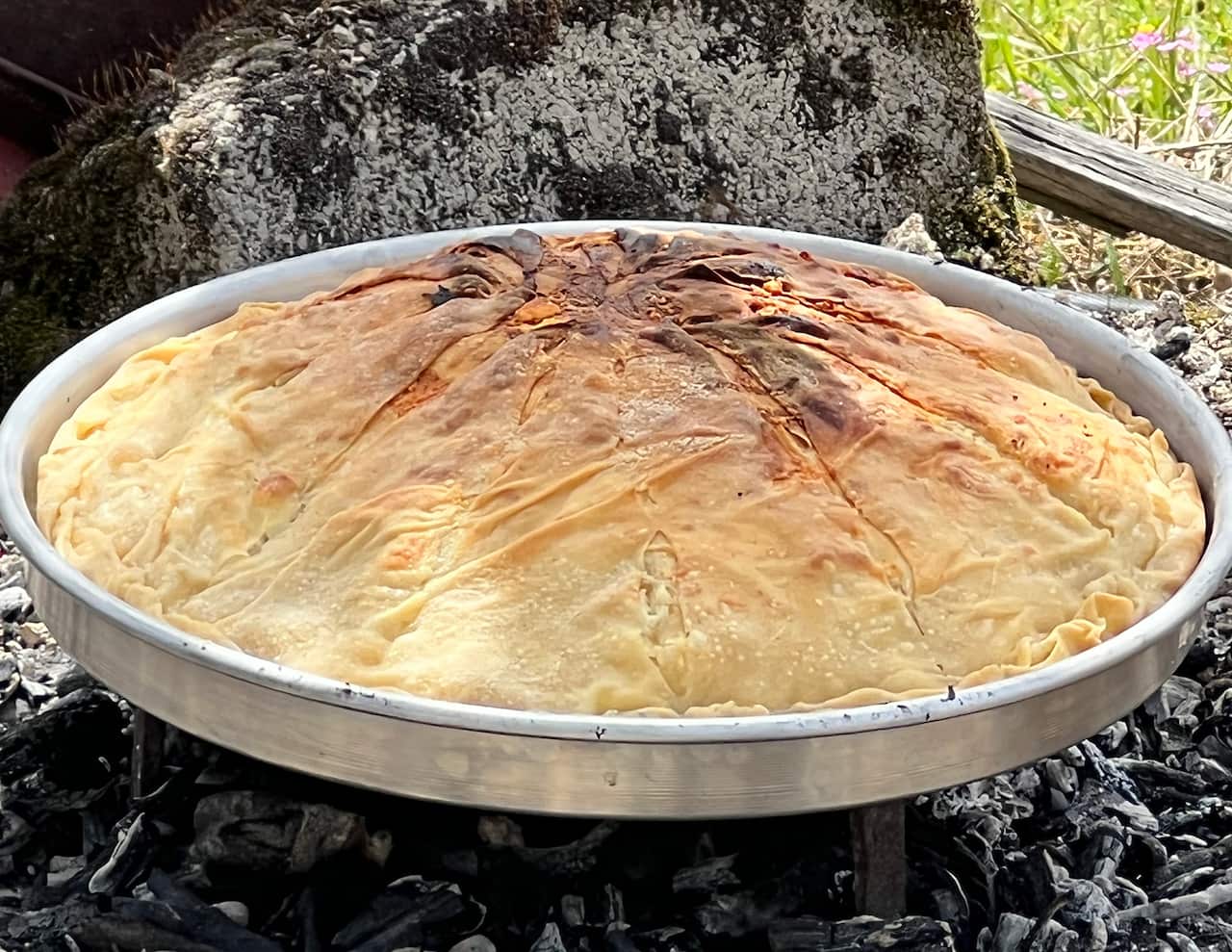 A large round silver pie tin with a cooked Greek pie in it sits above a pile of grey-black embers and ashes. 