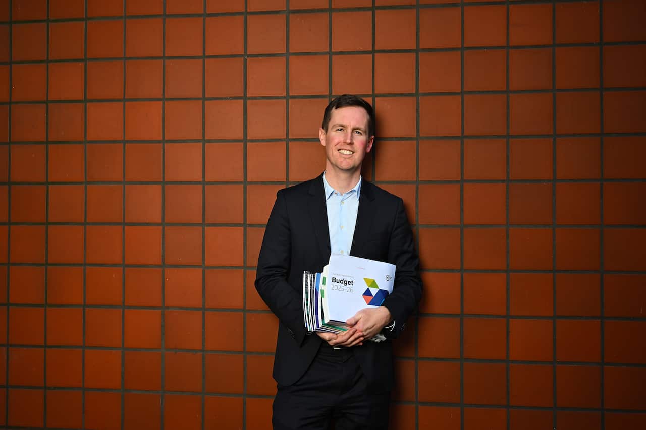 A man wearing a black suit and light blue shirt, standing in front of a dark red tile wall. He's holding a stack of papers that says "Budget".