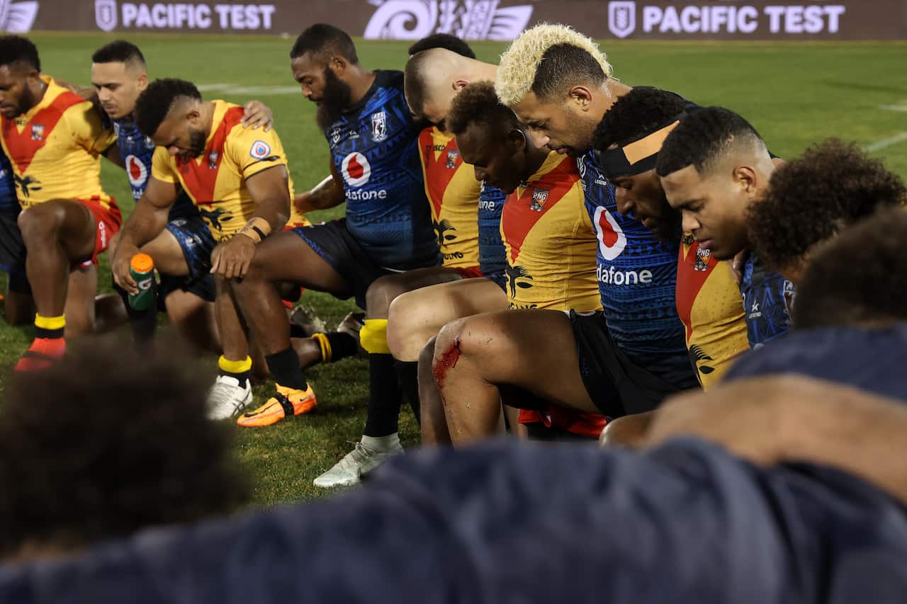 PNG and Fiji players kneel down on one knee with eyes closed looking down, praying, on the football field. 