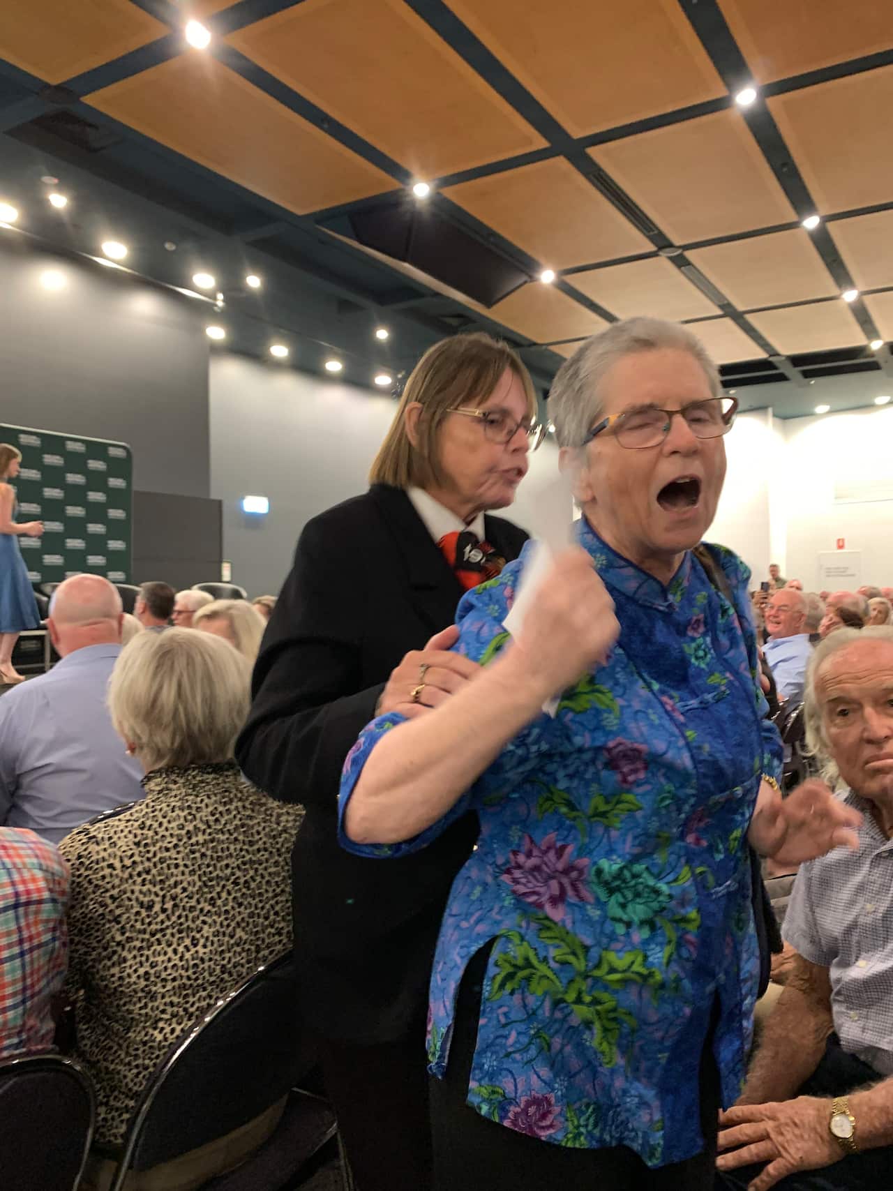 An elderly lady with her mouth open in distress with a security guard holding her arm