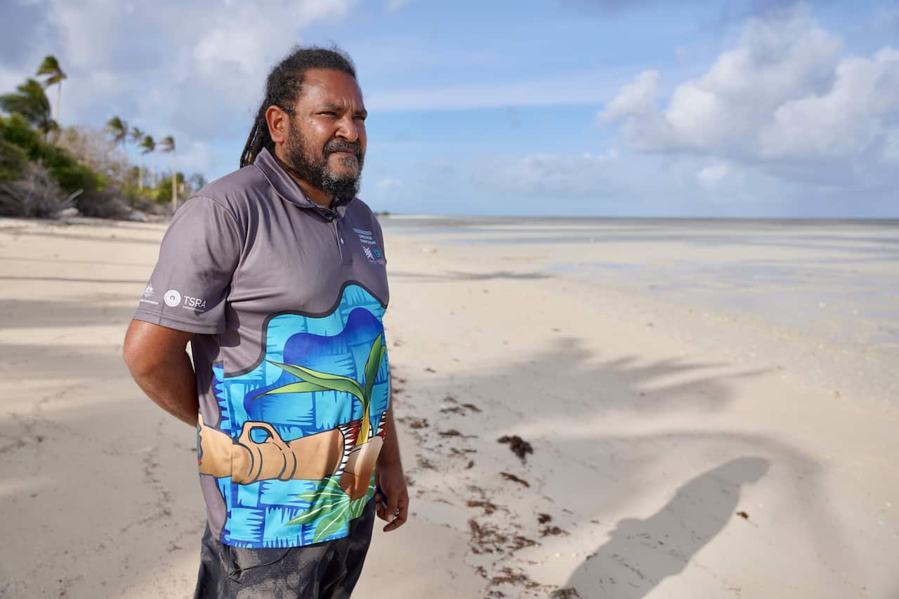 Yessie Mosby, a Kulkalgal man wearing a colourful shirt featuring a coconut palm, stands on the shoreline of Masig Island.