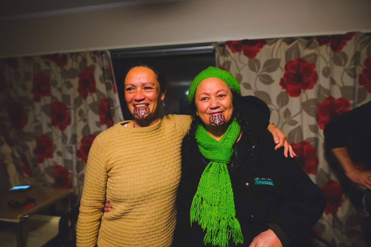 Two women smiling with traditional tattoos on their chins, standing side by side. One has her arm around the other.