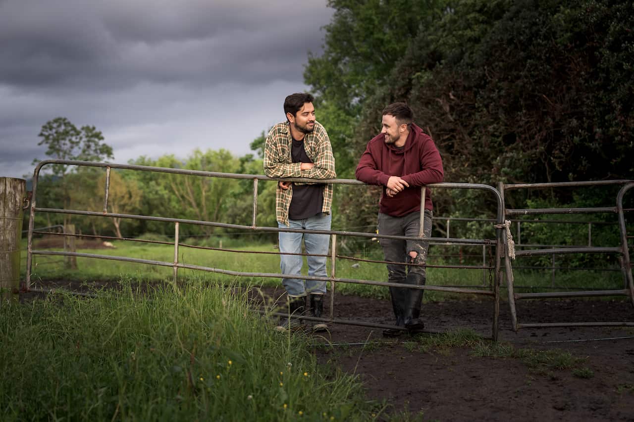 Two men lean on a sagging metal farm gate.