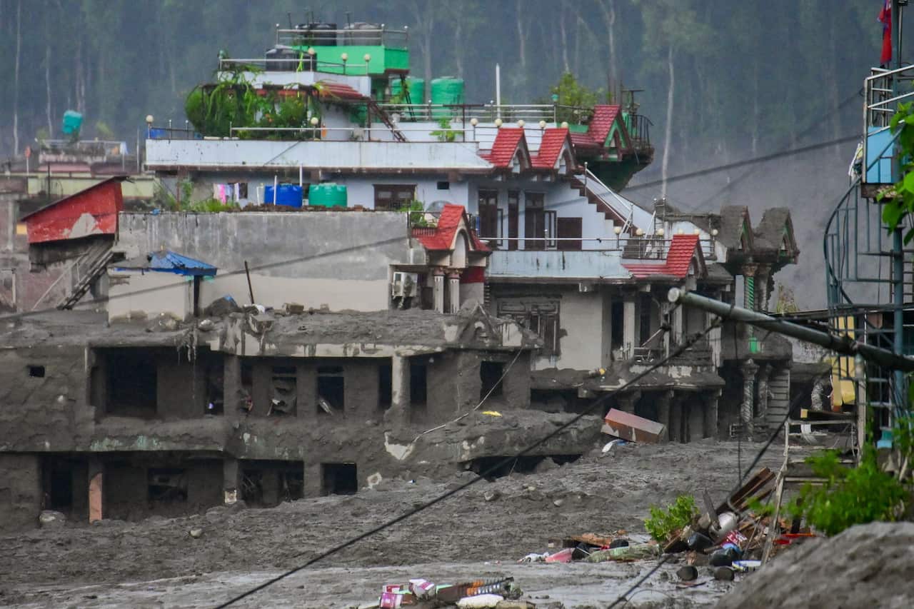 Buildings partially buried in thick grey mud and debris after a destructive mudflow, with damaged structures, scattered wreckage and a forested hillside in the background.