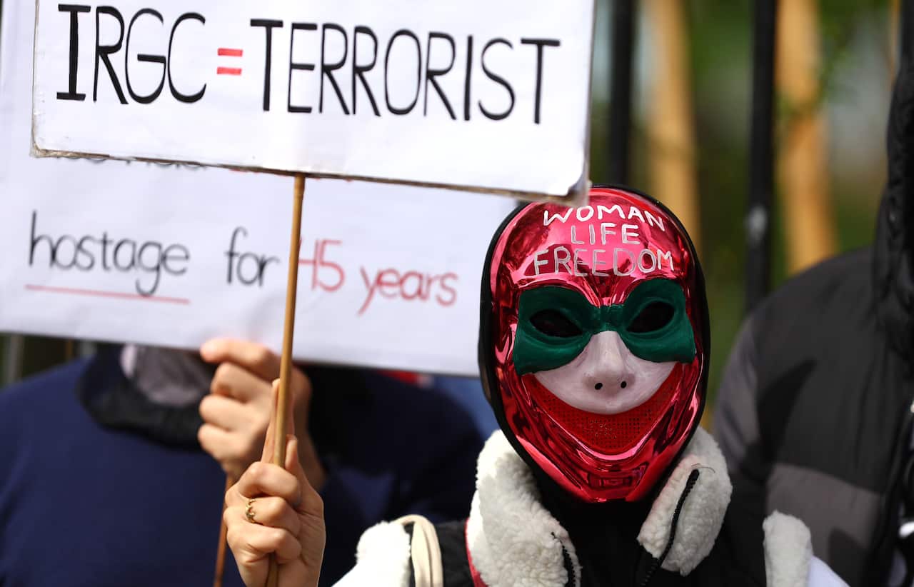 A person in a red metallic mask with the words Woman Life Freedom on it holds a sign reading IRGC=Terrorist