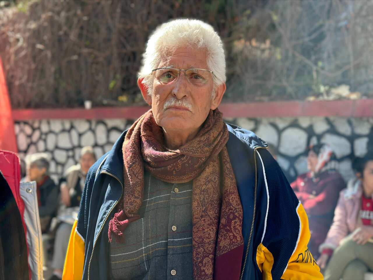 An old man wearting glasses, a navy blue and yellow jacket, and a patterned scarf