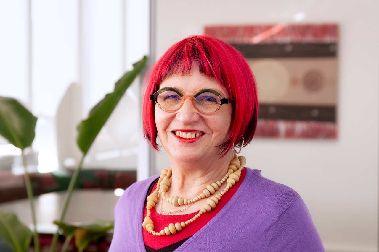 A woman with pinkish-red hair, chunky glasses, and a purple and red top