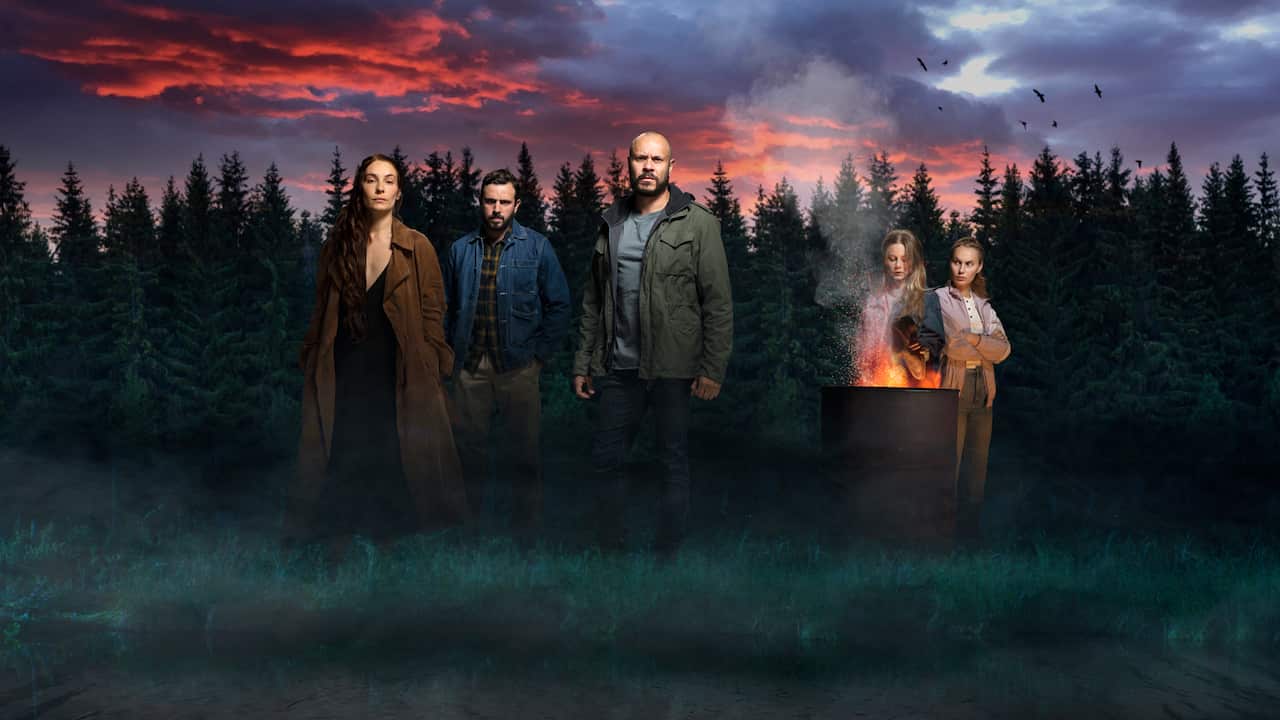 3 people stand in a forest with 2 standing in the background by a barrel fire