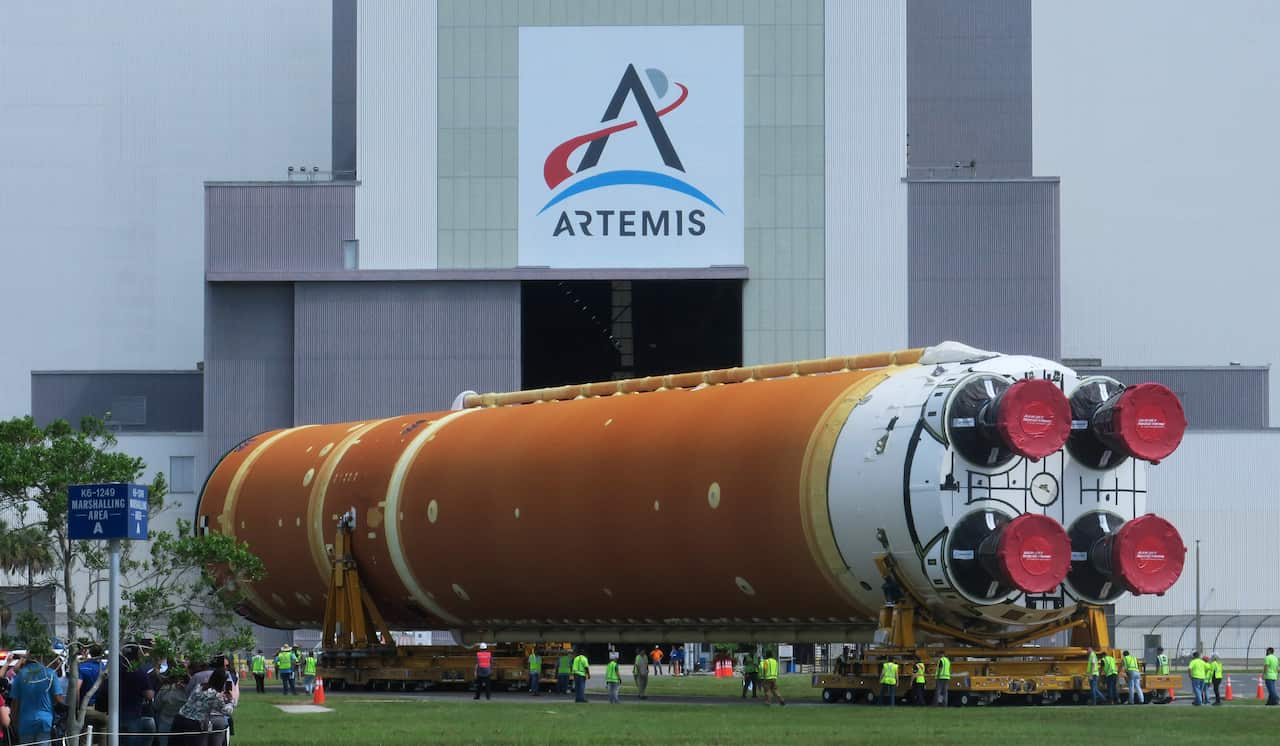 A massive, orange and white rocket core stage is being transported on a multi-wheeled vehicle in front of a large building with the "ARTEMIS" logo on it. A group of people in bright vests are walking alongside it, giving a sense of its immense scale.