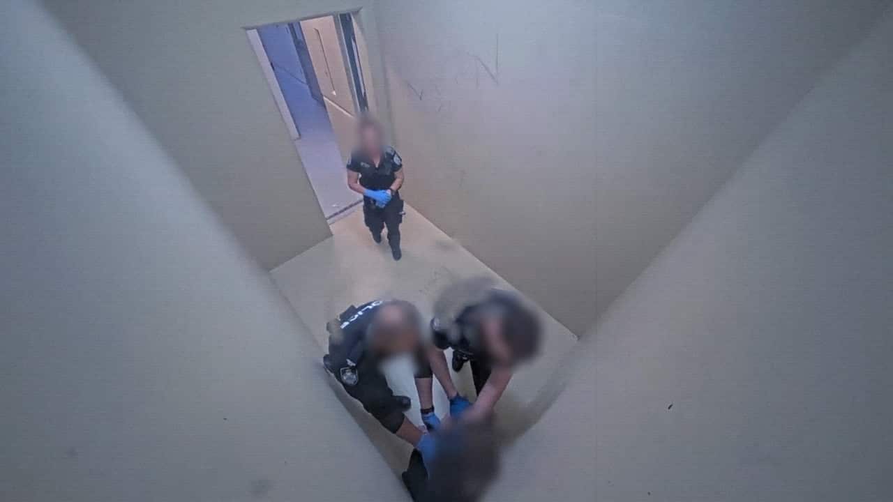 A teenage girl is pressed against the corner of a tiny, windowless cell by two uniformed officers. A third officer watches by the door.