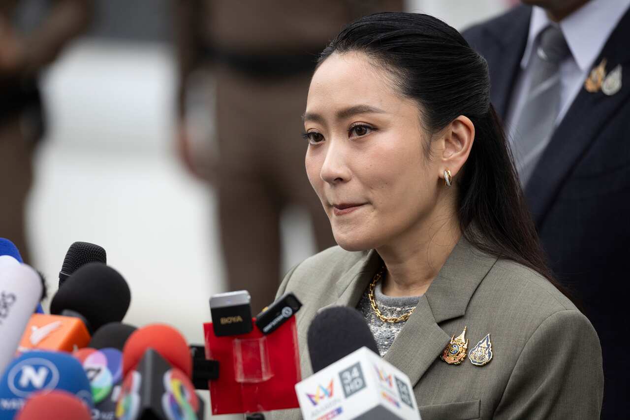 A woman wearing a grey blazer speaking into a large array of media microphones.