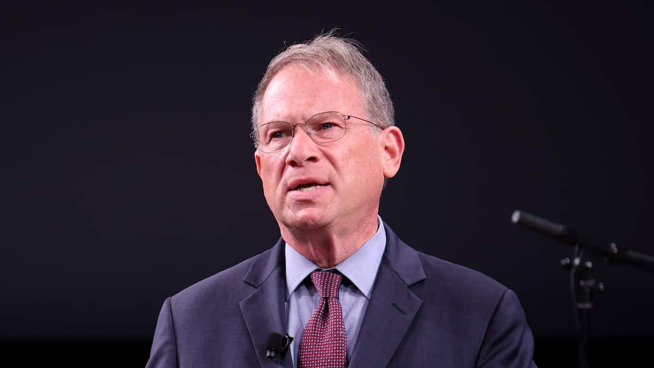 A man in a blue suit and maroon tie is speaking in front of a black background.