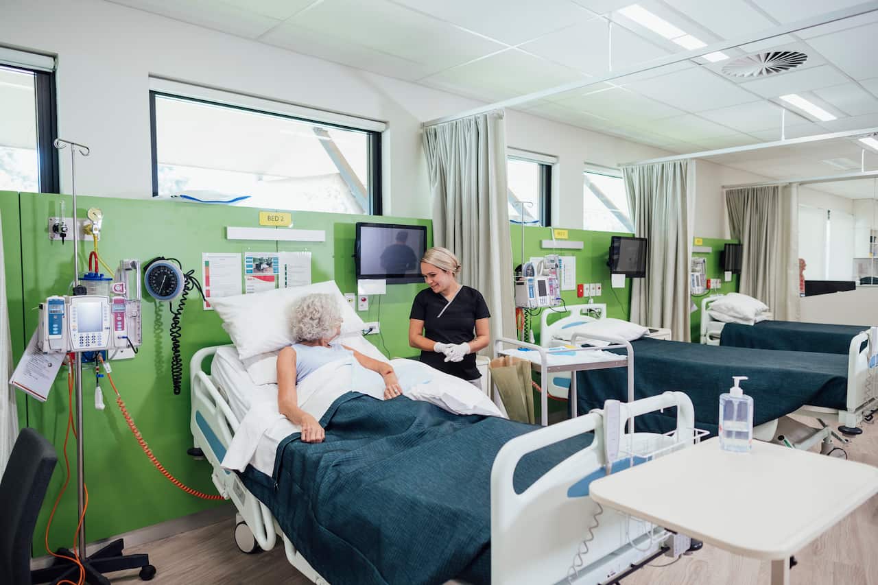 A nurse standing and speaking to a patient in a bed in a hospital ward 