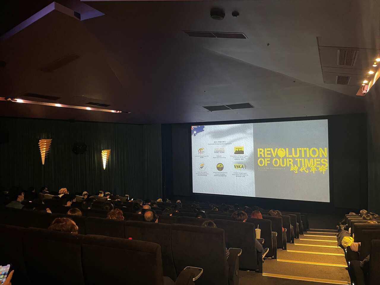 A cinema theatre full of people who are looking at a big screen that reads 'Revolution of our Times'.