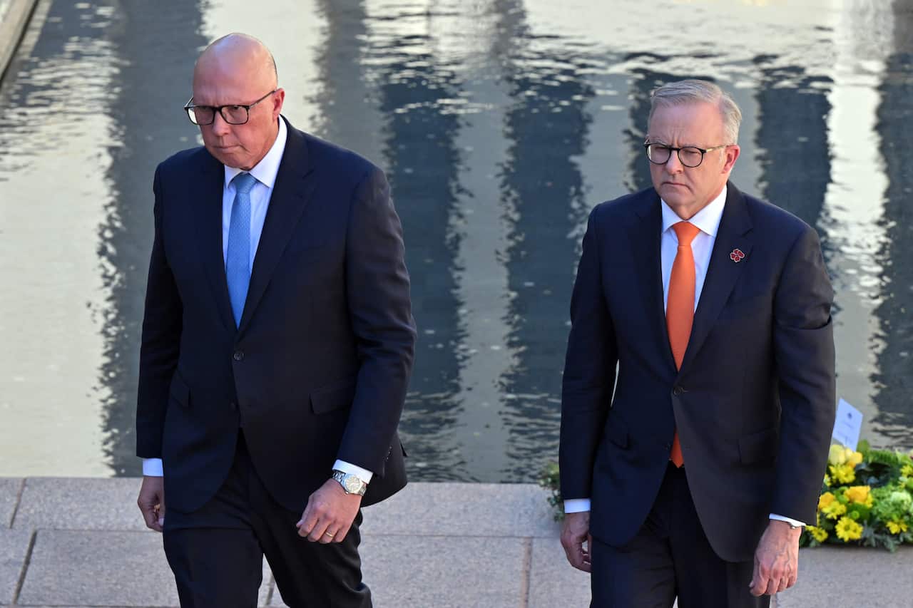 Two men in suits and ties, looking serious, with the water from a memorial site in the background.