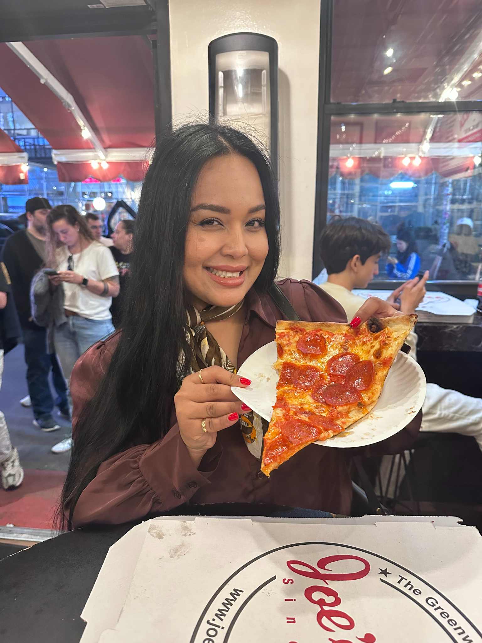 A woman with brown hair and brown eyes is holding a large slice of pizza while smiling