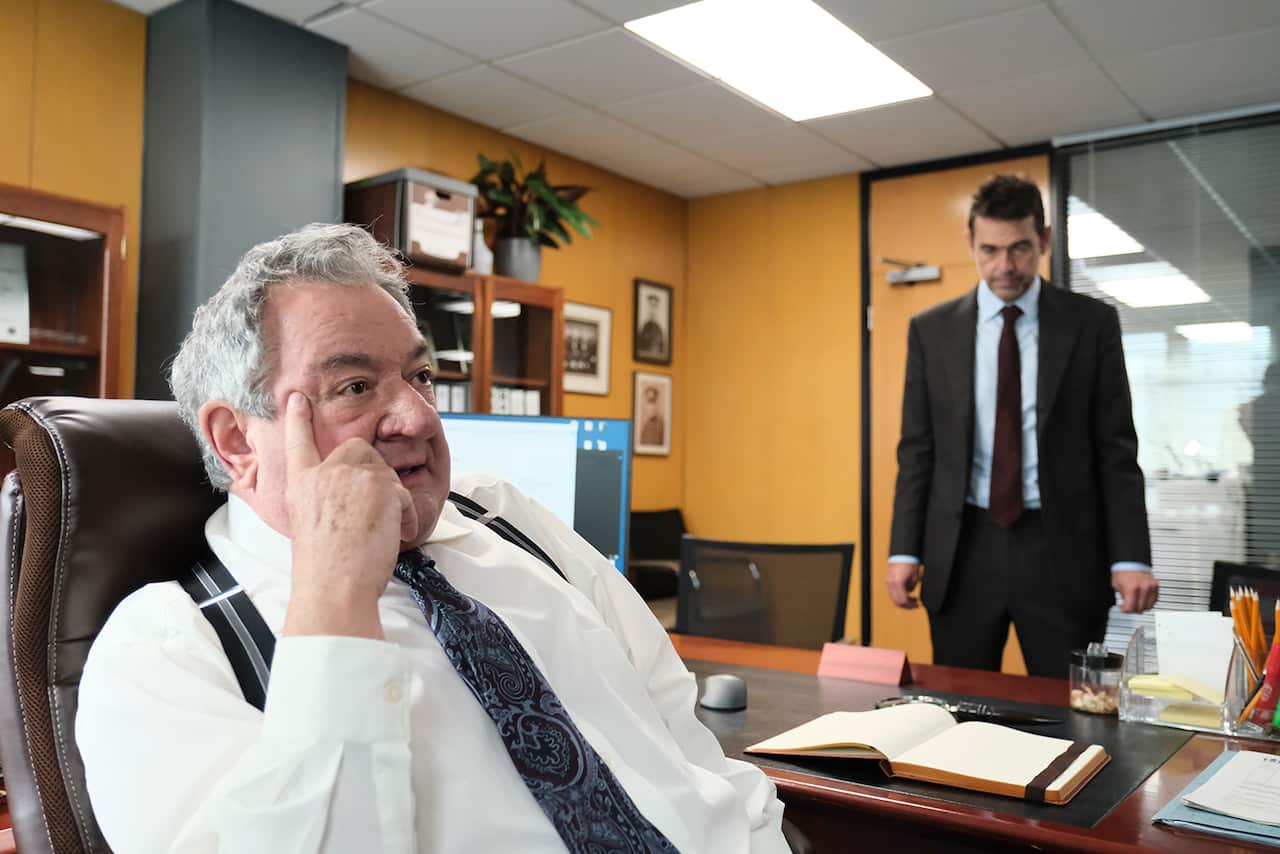 A grey-haired man, wearing a white shirt, suspenders and tie, is at the front of the image, sitting in a chair sideways on to a desk. On the far side of the desk, a man in a suit sands inside the door to the office.
