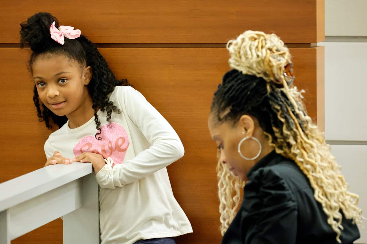 A young girl standing while her mother, looking down, is seated in a courtroom.