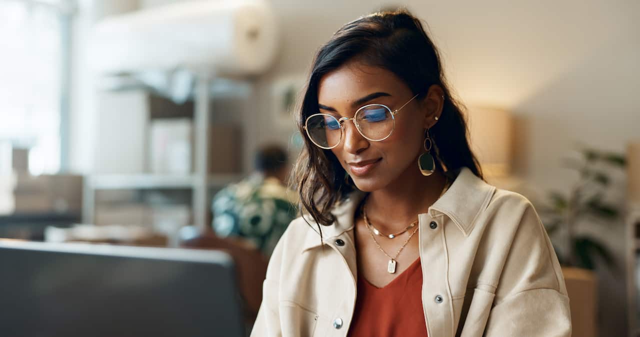 A woman with dark hair and glasses looks down at a laptop.