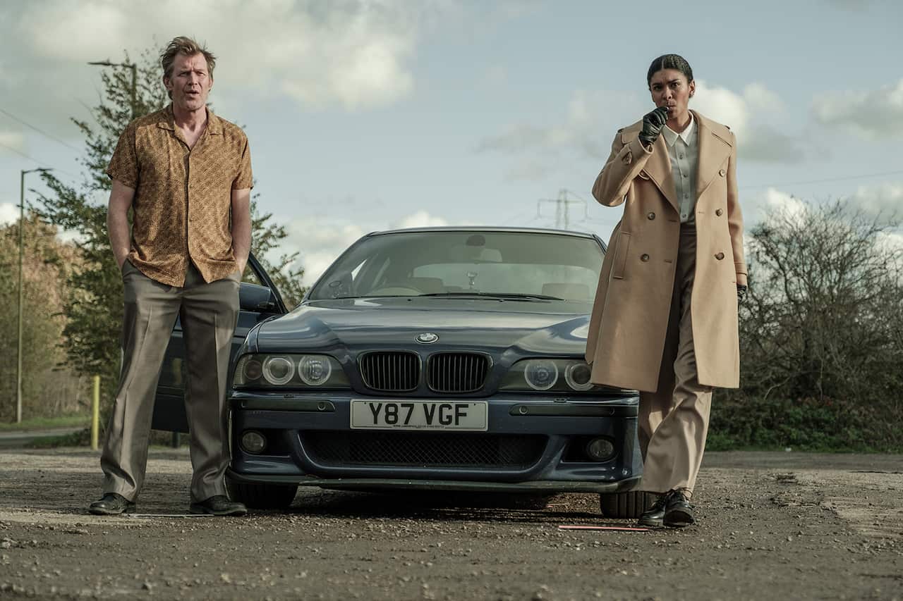 A man with messy hair and crumpled shirt stands on one side of a BMW. An elegantly dressed woman in a tan coat and black gloves stands on the other side.