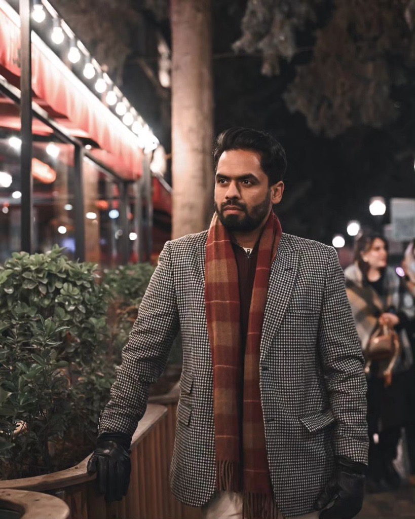 A man in a grey blazer and a red-and-grey checked scarf stands outside a well-lit restaurant. 
