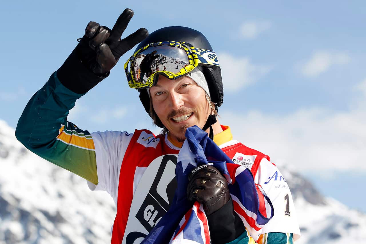 A man in snowboarding gear smiles. There is snow in the background