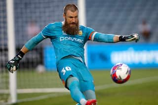 A bearded goalkeeper in a teal jersey and shorts kicks a ball down the field during a match.