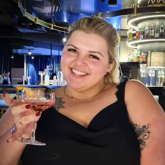 A blonde woman with tattoos wearing a black top and holding a cocktail